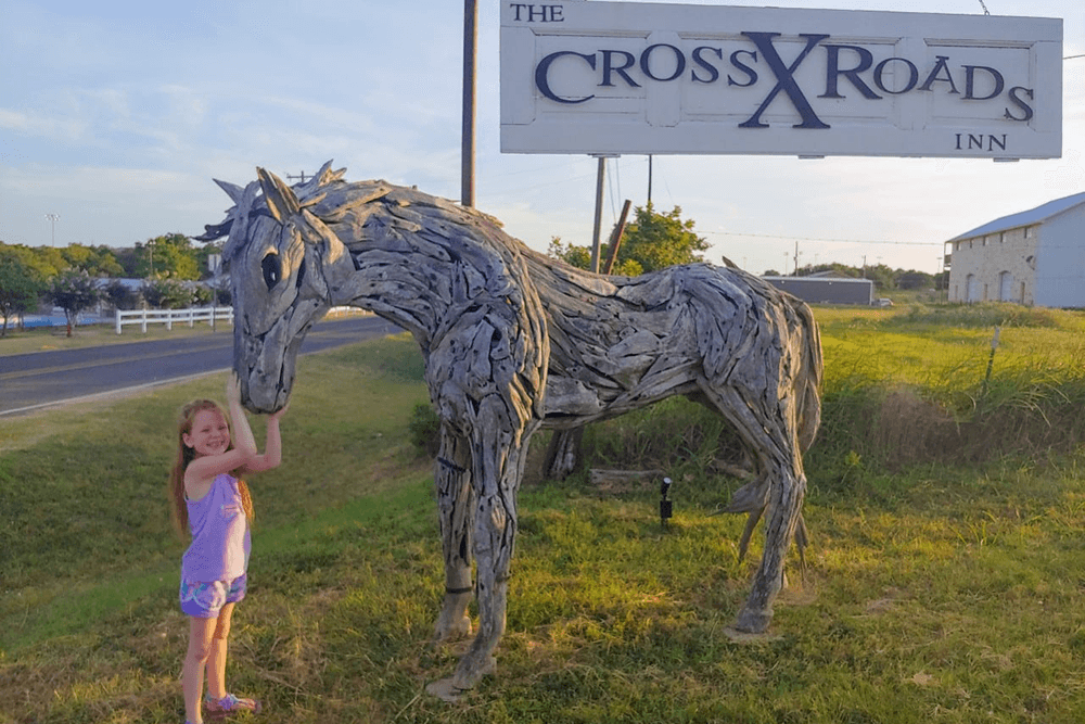 A young girl smiles while touching a large wooden horse sculpture next to the Crossroads Inn sign.