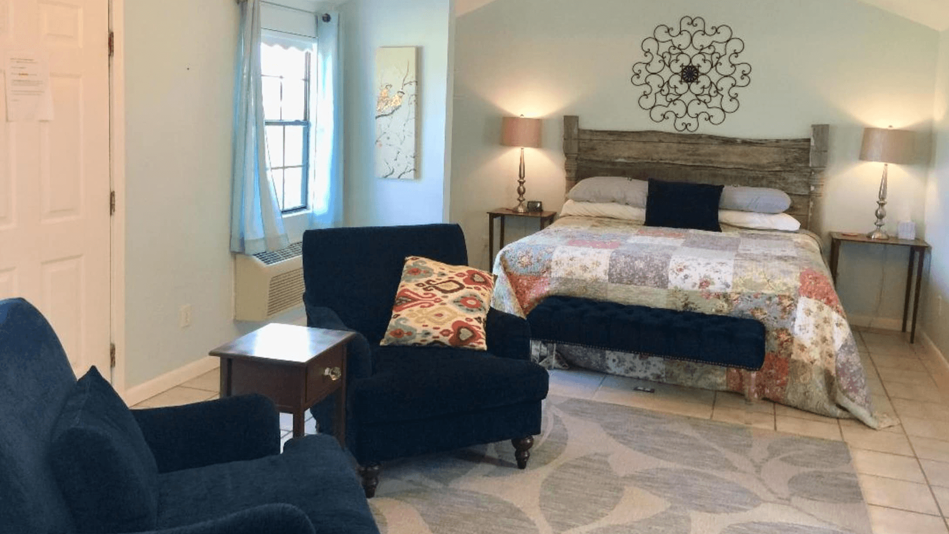 A cozy bedroom featuring a quilted bedspread, armchairs, and soft lighting.