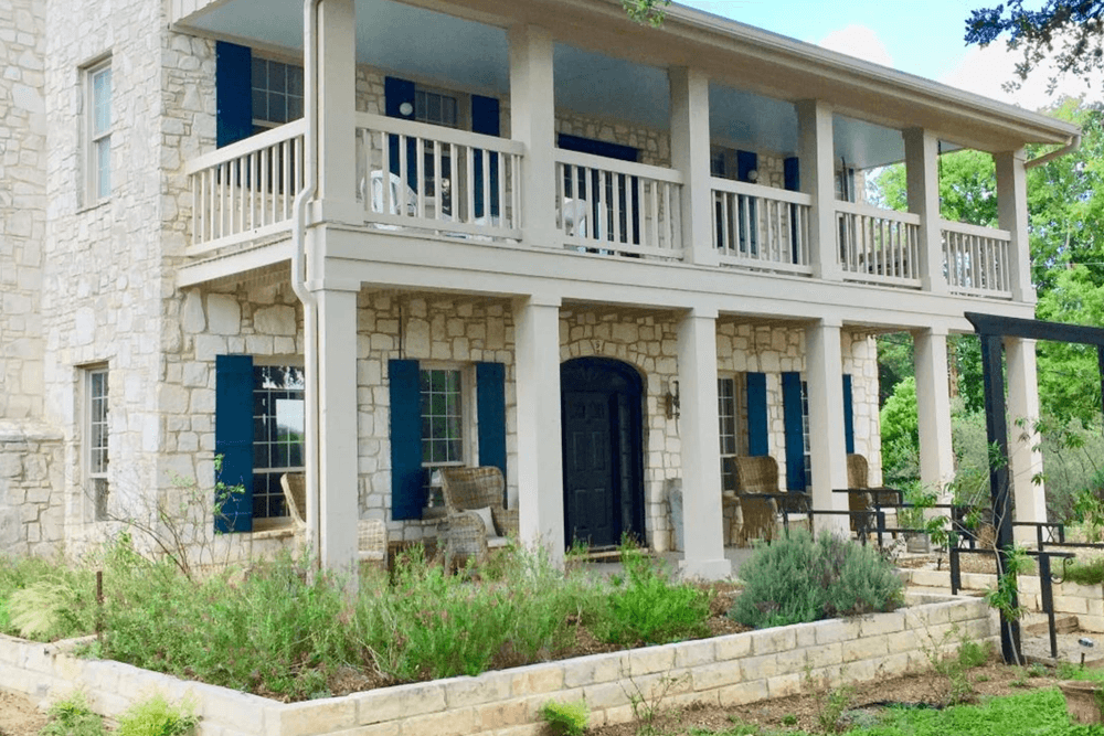 Two-story stone house with blue shutters and a front porch, surrounded by greenery.