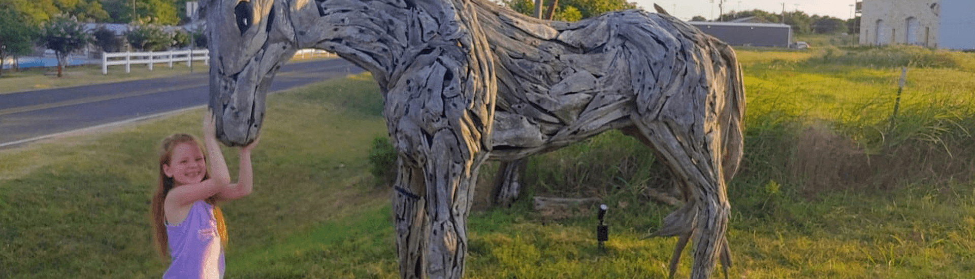 A girl poses beside a wooden horse sculpture in a grassy area.