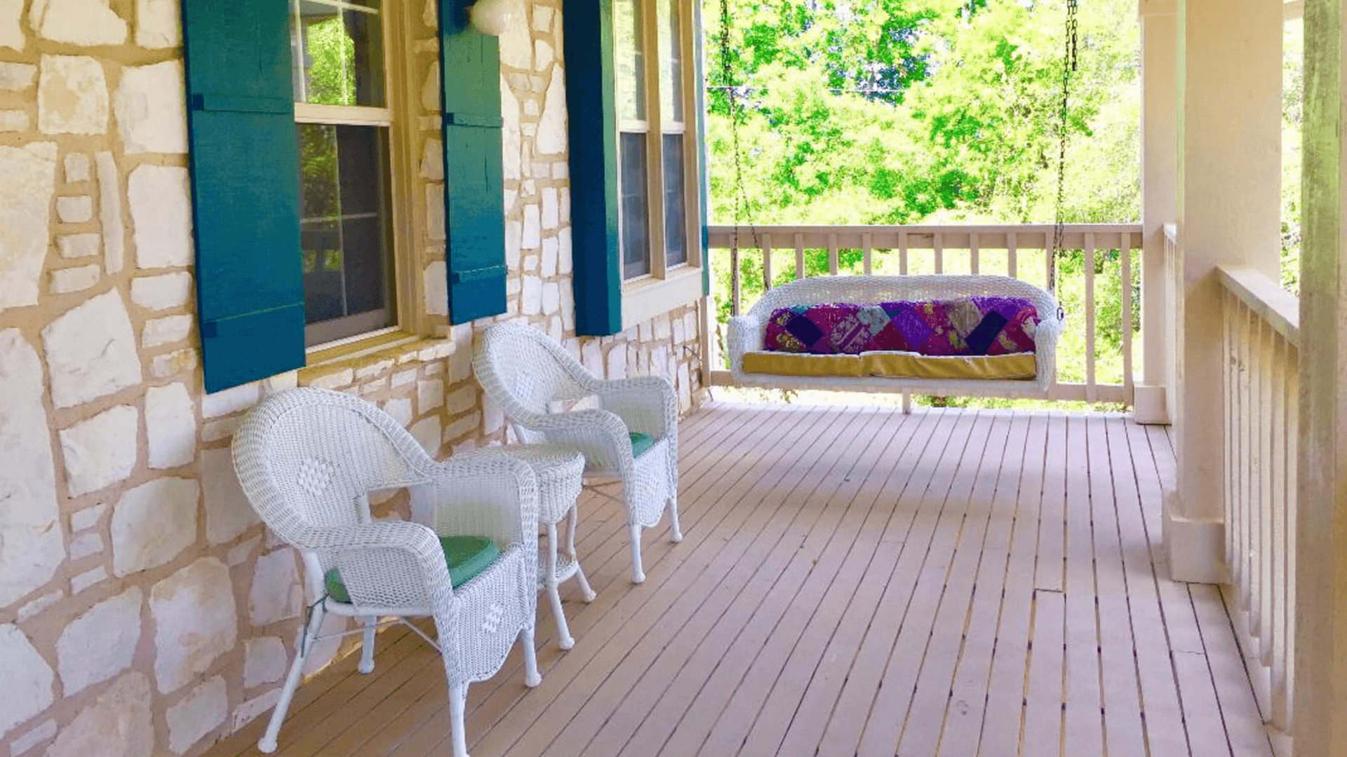 A cozy porch featuring two white wicker chairs and a hanging swing, surrounded by greenery.